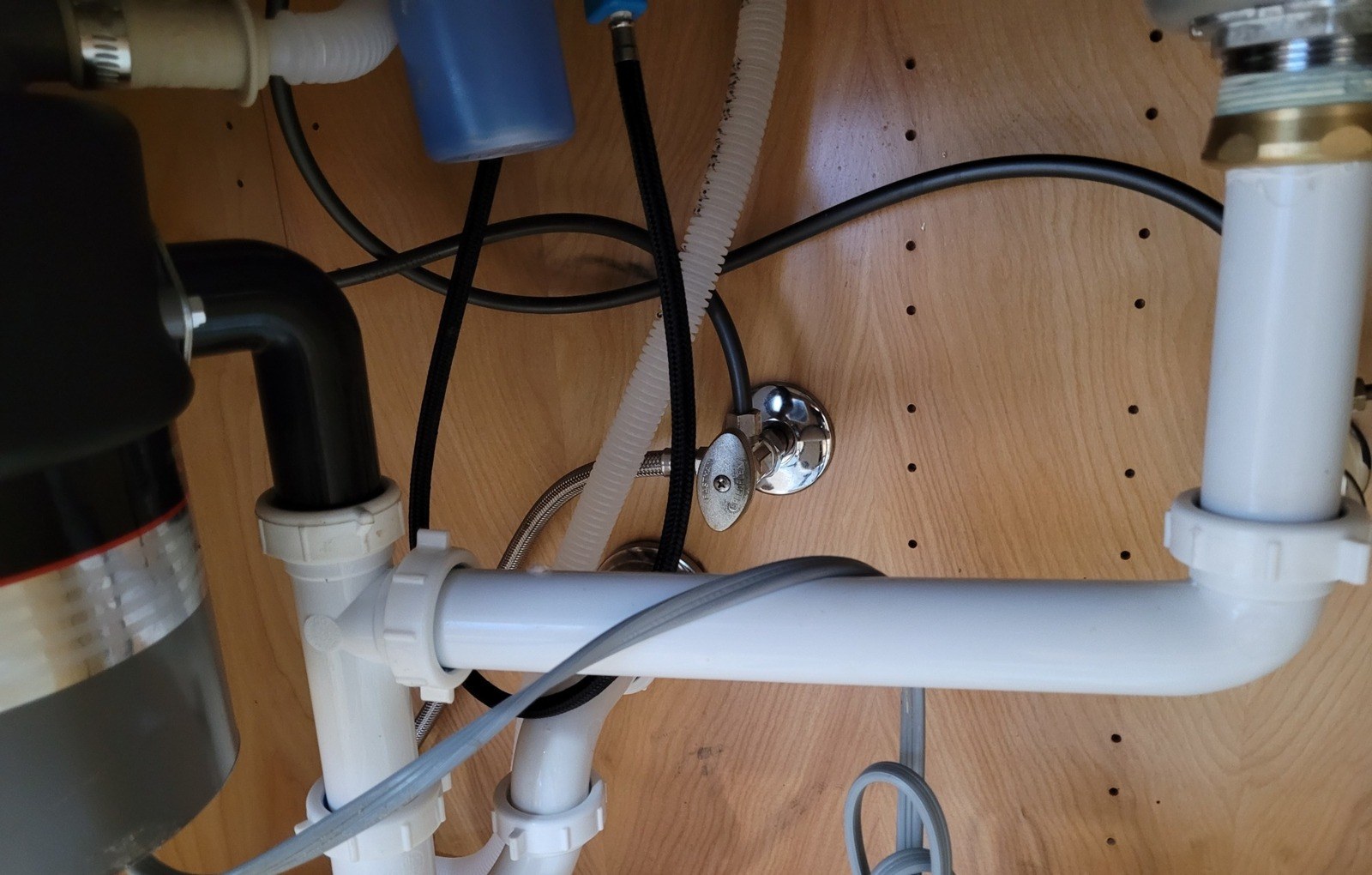 Under a kitchen sink, a new chrome double angle stop feeding the faucet and the dishwasher, beside a white plastic P-trap and a black disposal body on a wood cabinet back