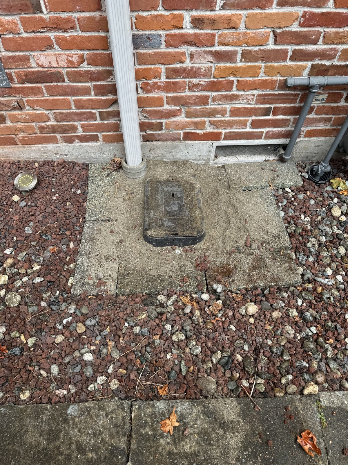 Square black valve box lid set into red lava rock and gravel against a brick wall, beside a white downspout
