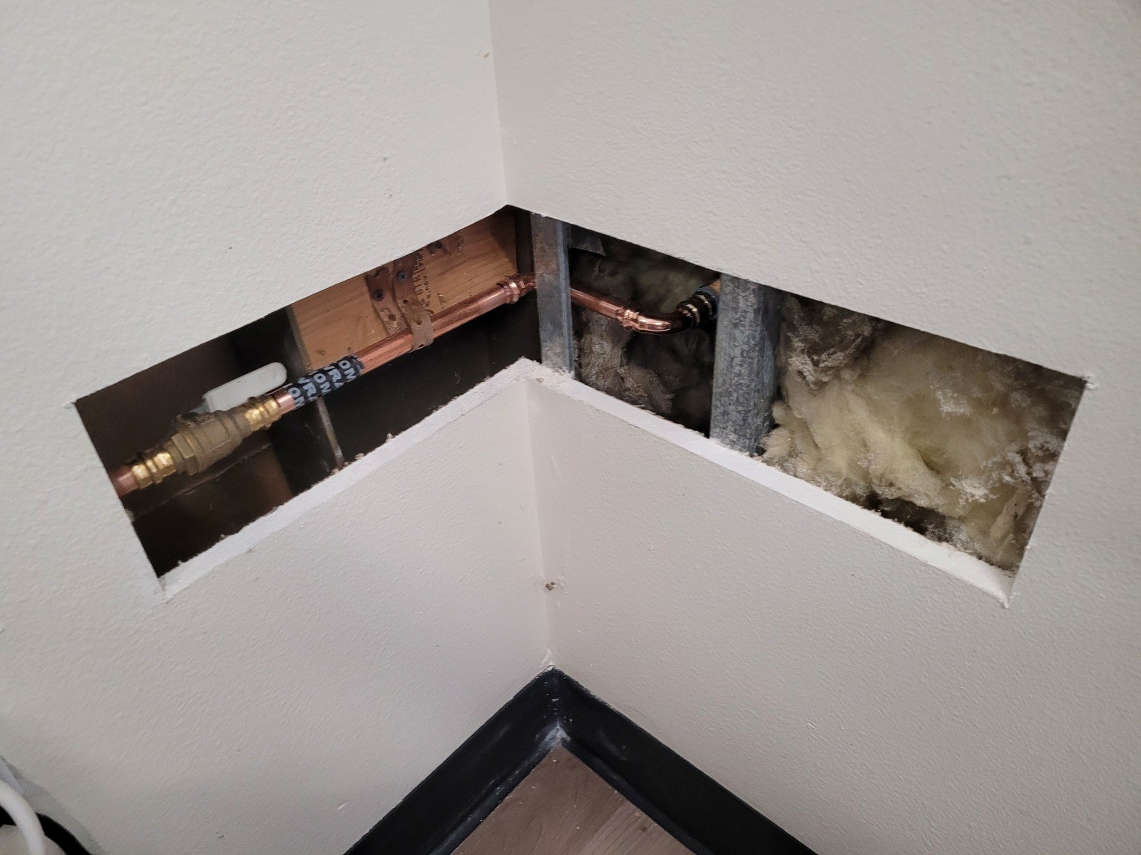 Opened drywall at an inside corner showing new copper pipe with a brass ball valve, a push-fit coupling and insulation in the wall cavity above a black baseboard
