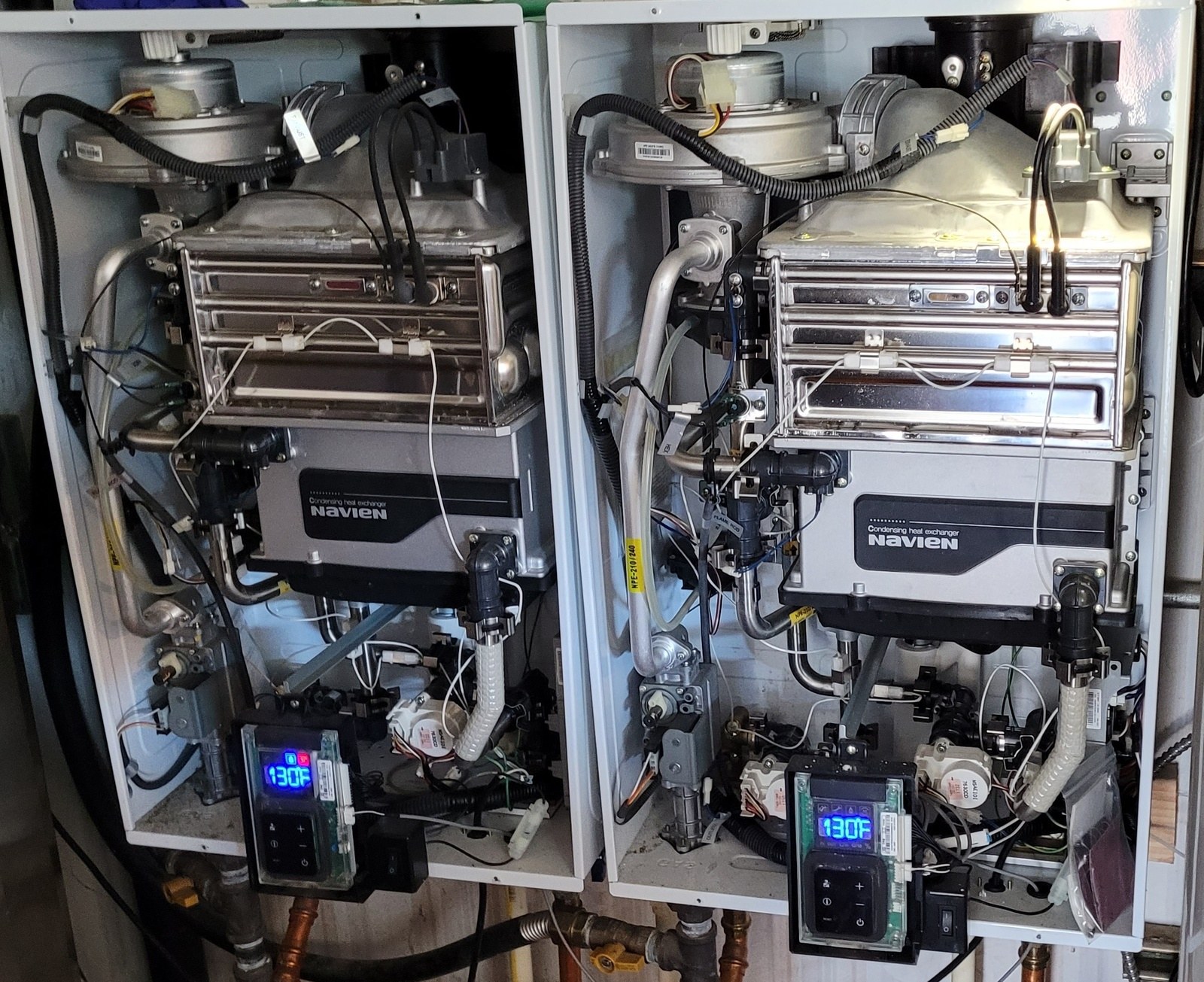 Two white Navien tankless water heaters mounted side by side with their covers off, both displays reading 130 degrees, above a copper manifold