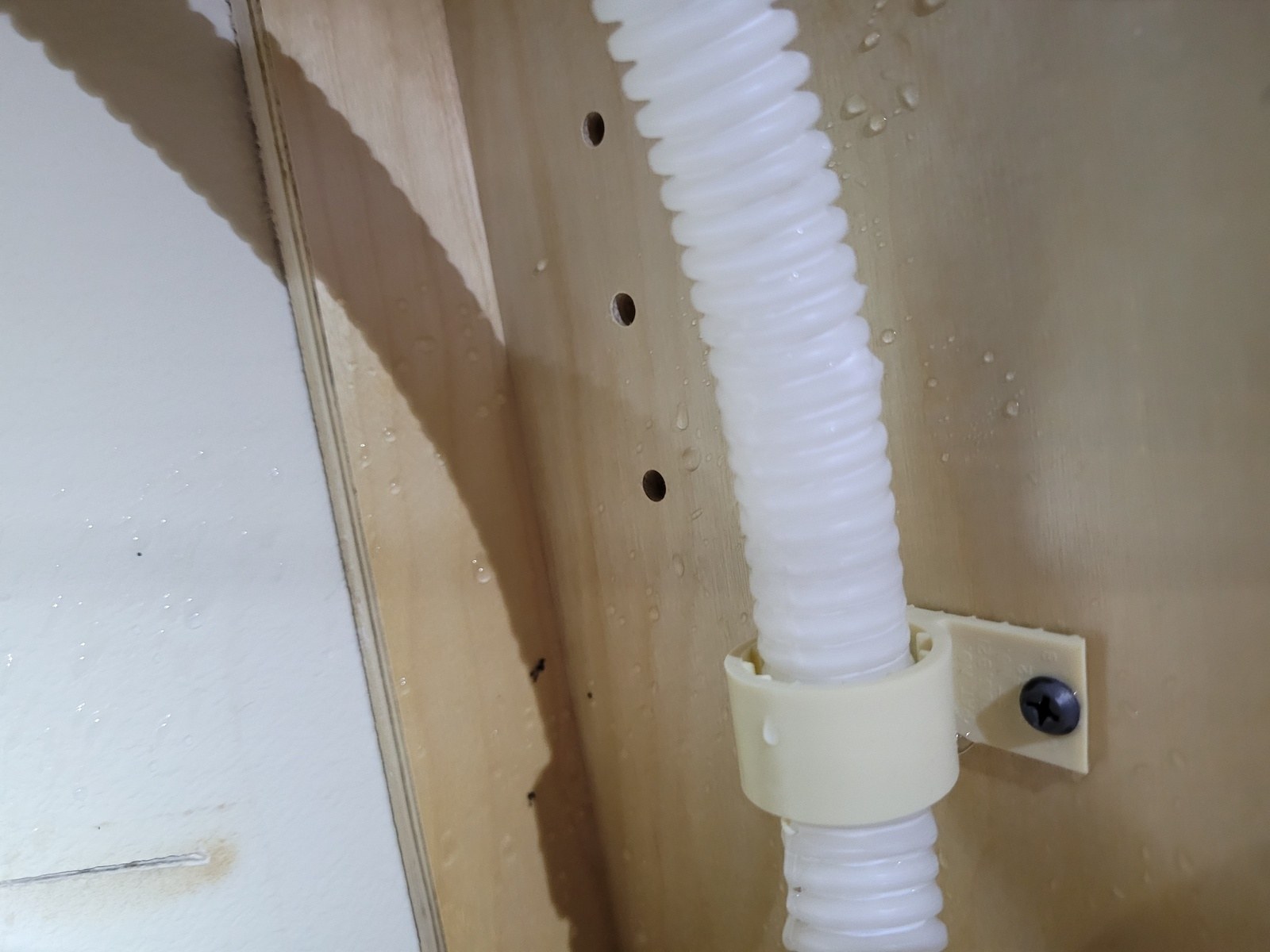 Corrugated dishwasher drain hose clipped inside a cabinet wall with water droplets visible around it