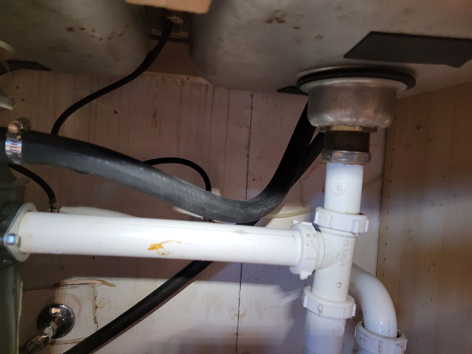 White PVC kitchen sink drain with a disposer outlet and a black dishwasher discharge hose under a stainless sink, on a plywood cabinet wall