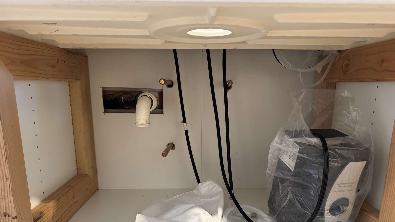 Open sink cabinet before the trim-out, with copper water stubs and a white drain stub coming through the wall, the new faucet's hoses hanging down and the instant-hot tank still in its plastic bag