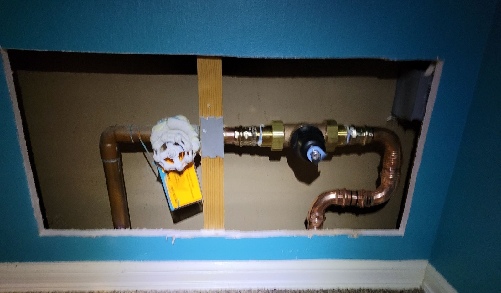 Copper water main inside an opened closet wall, with a white-handled gate valve, a yellow manufacturer’s tag and a new black-capped pressure-reducing valve on press fittings