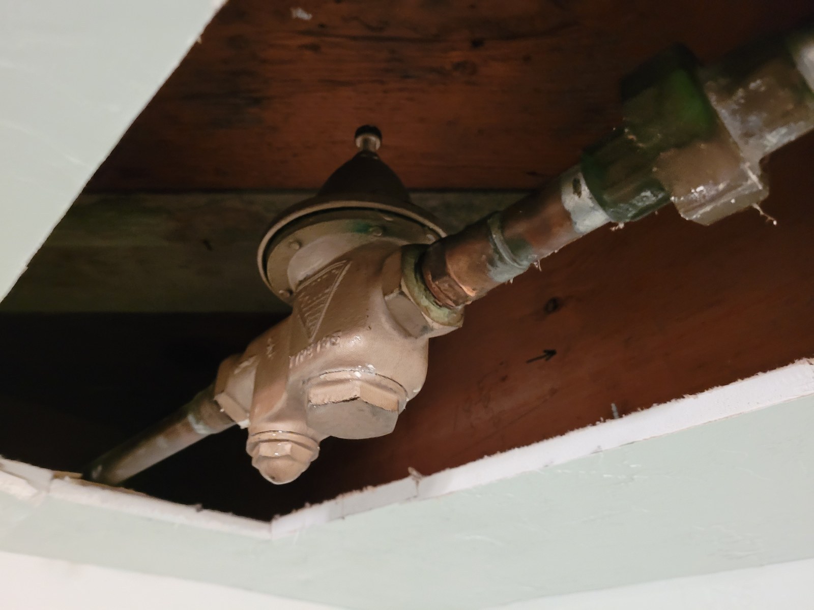 Old bronze pressure-reducing valve with green corrosion on its fittings, angled across an opening in a basement ceiling