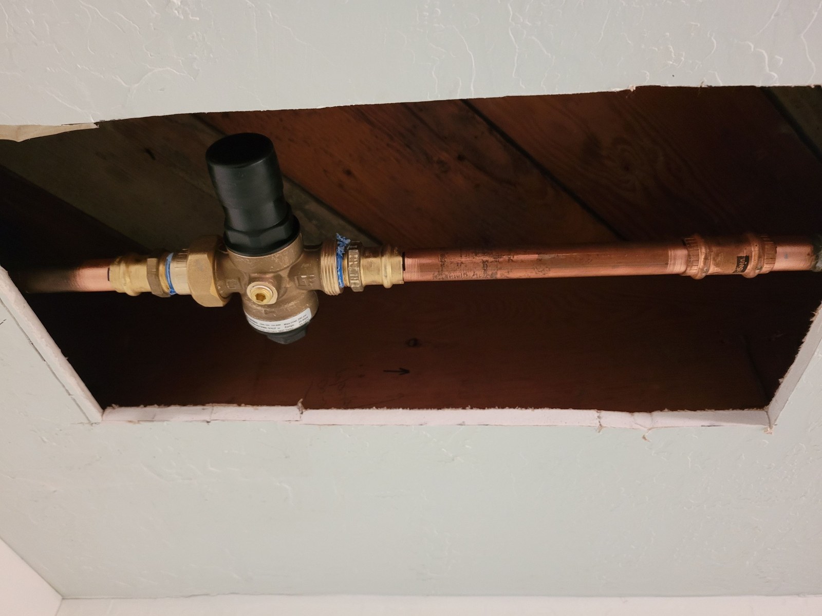 New brass pressure-reducing valve with a black bell on new copper pipe, seen through a square opening in a white basement ceiling