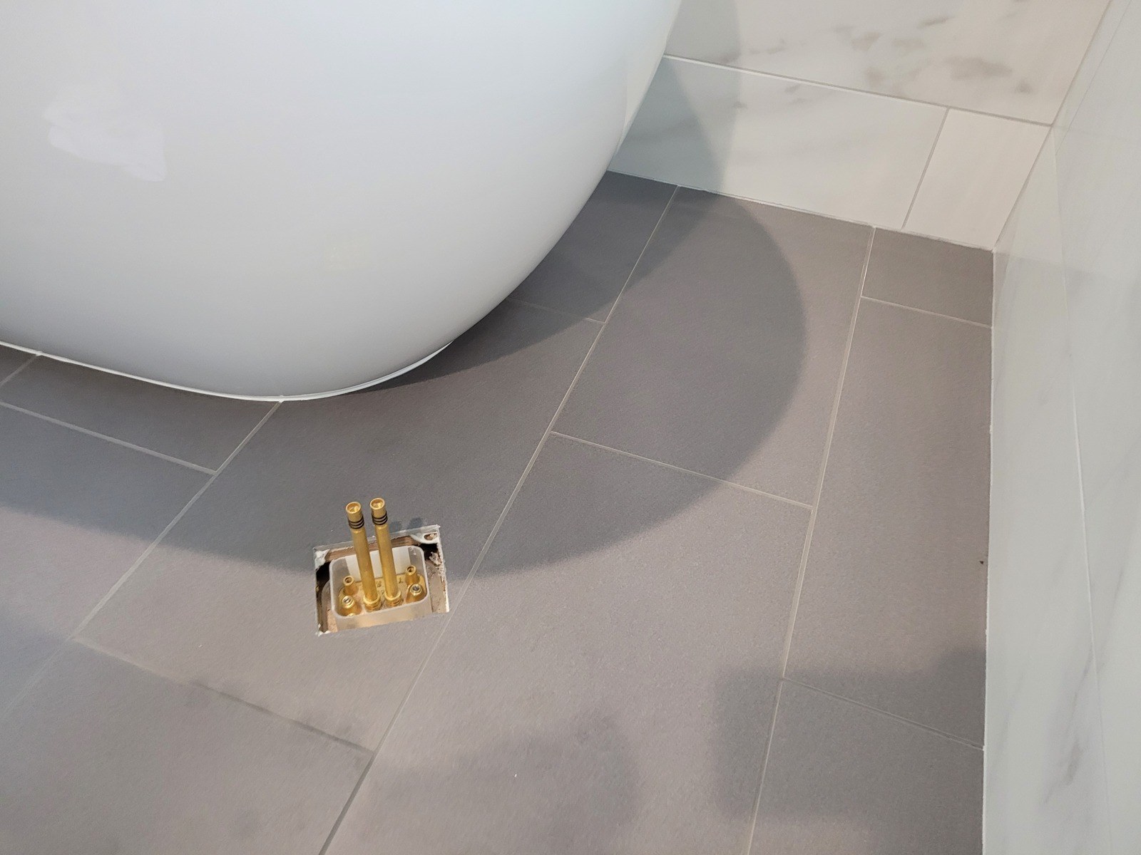 White freestanding soaker tub on grey floor tile beside a marble-look tiled wall, with a brass floor rough-in box and two supply stubs set flush in the tile