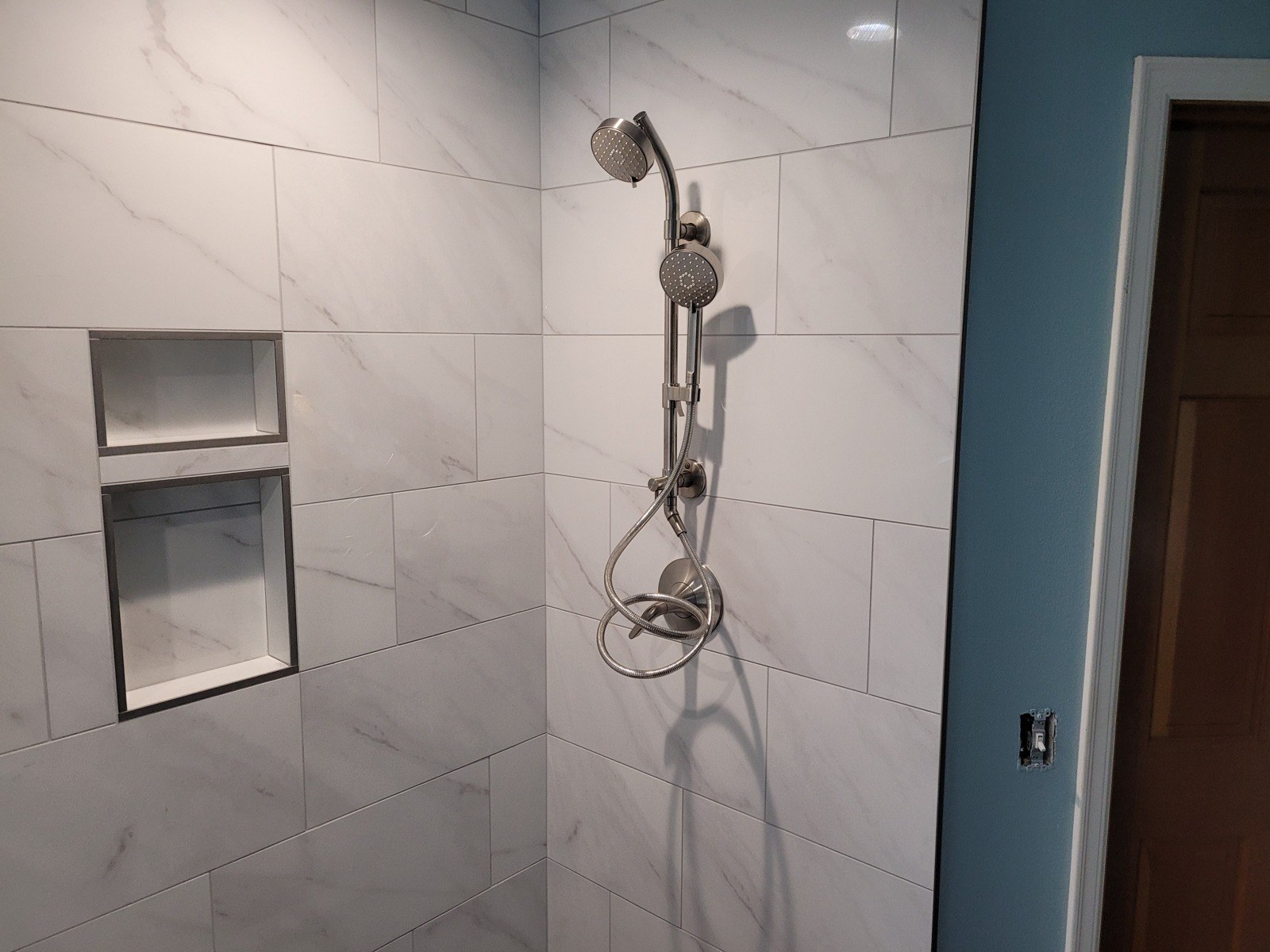 Brushed-nickel shower slide bar with a fixed head and a handheld sprayer on white marble-look wall tile, beside a two-shelf recessed tile niche
