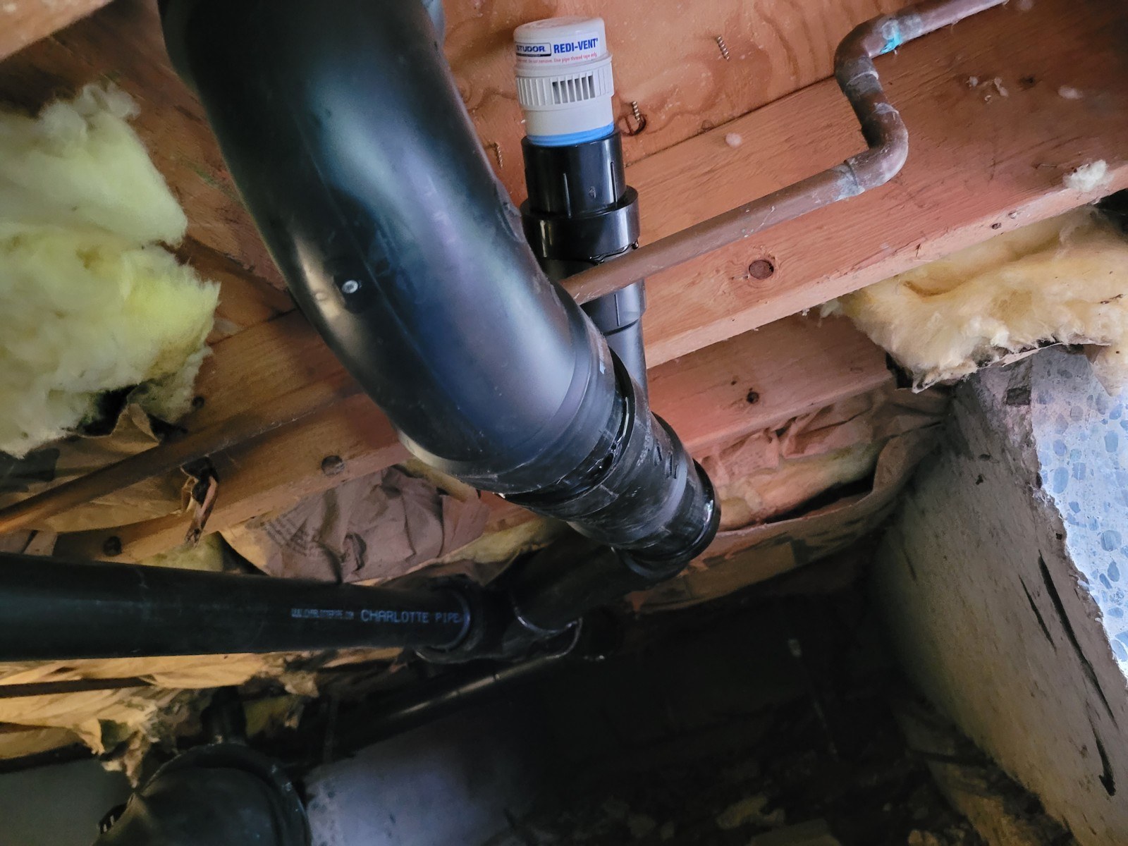New black ABS toilet drain under a floor in a crawl space, sloping down to a wye, with a white mechanical vent standing up from the line and pink insulation around the joists